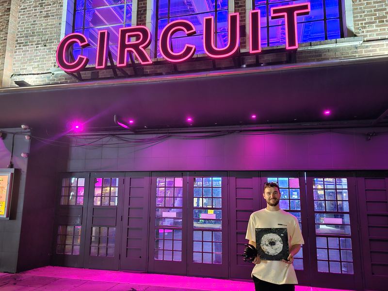 Dindalé holding a record outside Circuit under its pink neon sign